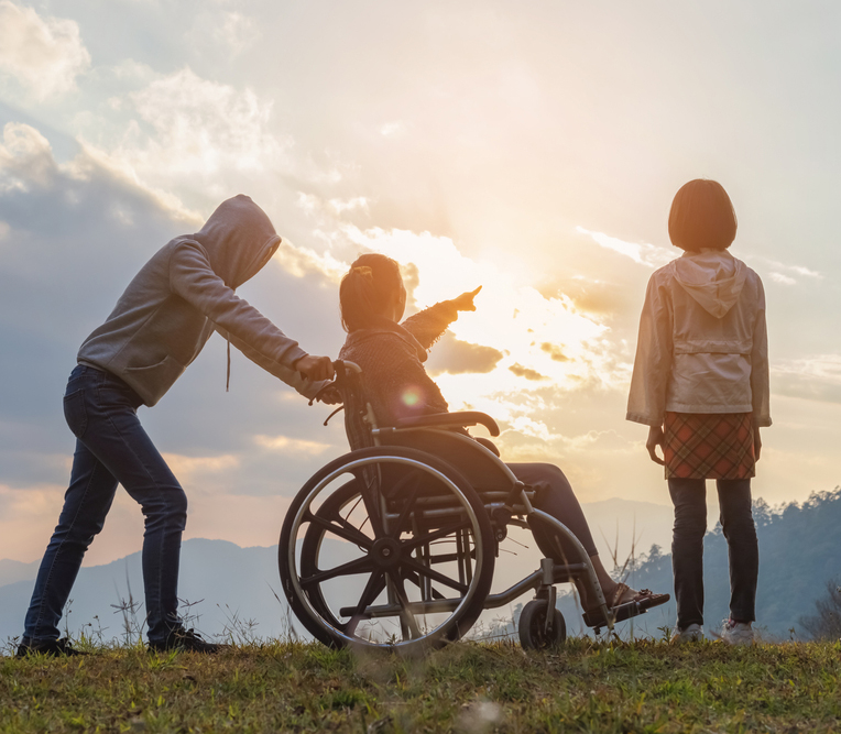 Paralyzed woman sitting on her wheelchair with her daughter and care helper at sunset